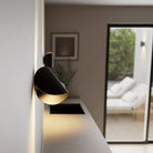 Modern black wall sconce illuminating a minimalist living room with natural light, featuring a large window and neutral decor.