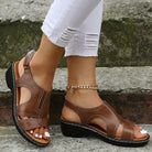 Women's brown leather sandals with wedge heel, featuring cut-out design and ankle strap, paired with white ripped jeans. Fashionable summer footwear.