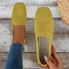 Yellow slip-on knit loafers with textured design, held by a hand over rustic wooden floor. Comfortable, stylish women's footwear, perfect for casual wear.