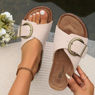 Beige women's sandals with large buckles, worn by a person with manicured nails, displayed outdoors on a textured surface. Fashionable summer footwear.