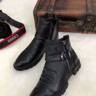 Black leather ankle boots with buckle and zipper detail on a white surface, next to a vintage brown suitcase and a Canon camera strap.