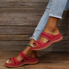 Red women's sandals with intricate cutout design, worn with ripped jeans, on a wooden floor. Stylish summer footwear, casual fashion, comfortable slides.