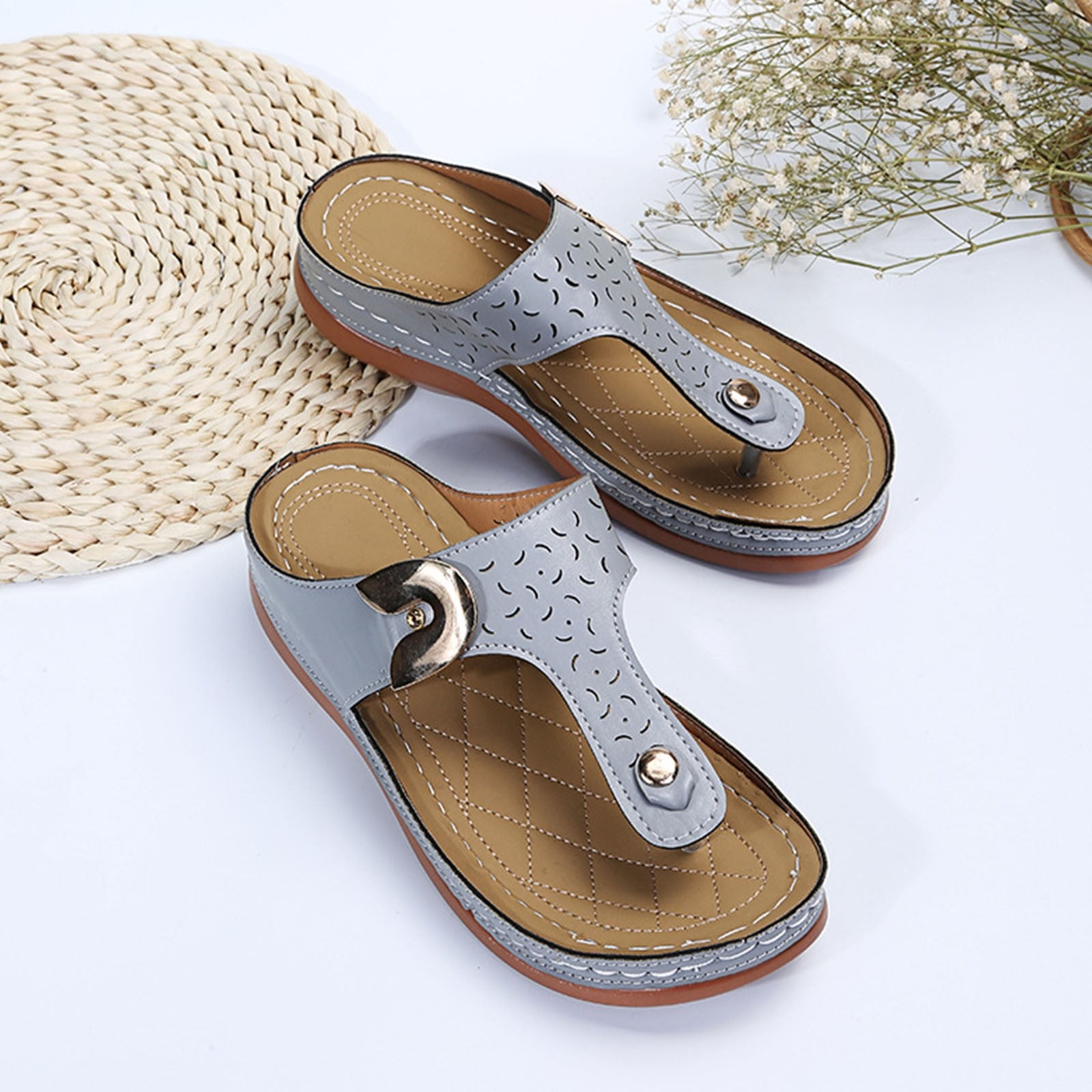 Gray leather women's sandals with decorative cutouts and cushioned soles on a white background, next to a woven mat and dried flowers.