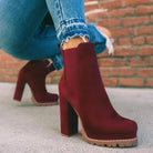 Women's burgundy high heel ankle boots with chunky sole, paired with distressed blue jeans. Stylish fall fashion footwear against a brick wall backdrop.