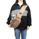 Person wearing a black and white checkered sweater and jeans, carrying a small dog in a tan pet sling carrier.