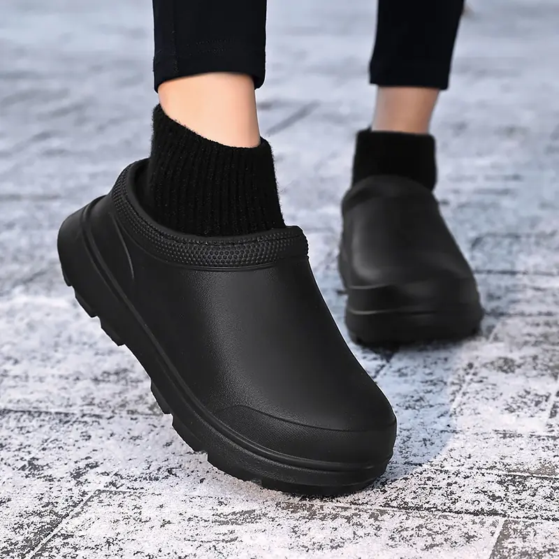 Black slip-on waterproof clogs with thick soles, worn with black socks on a textured gray pavement. Ideal for outdoor, casual, and rainy weather use.