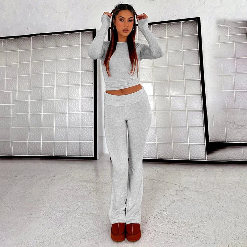 Woman wearing a gray two-piece lounge set with long sleeves and flared pants, standing indoors. Fashionable casual wear, comfortable home outfit.