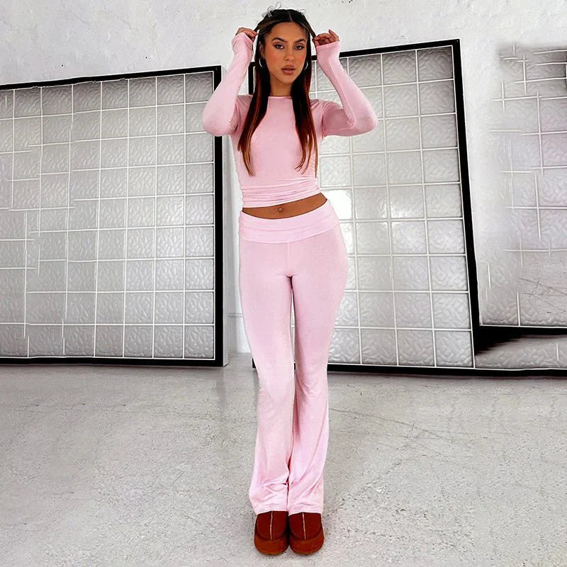 Woman wearing pink two-piece lounge set with long sleeves and flared pants, standing in a modern room. Fashionable casual wear, comfortable style.