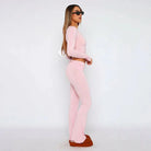 Woman in pink two-piece lounge set with long sleeves and flared pants, wearing sunglasses and brown shoes, standing against a white background.