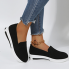 Black slip-on sneakers with white soles, worn with frayed hem blue jeans and a gold heart anklet. Stylish casual footwear for women.