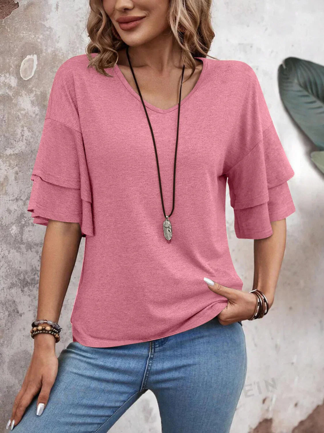 Woman wearing a pink ruffle sleeve top and blue jeans, accessorized with a long necklace and bracelets, standing against a textured wall. Fashionable casual outfit.