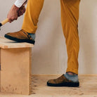 Person in mustard pants and brown shoes using a hammer on a wooden surface. Carpentry, DIY, woodworking, home improvement, casual footwear.