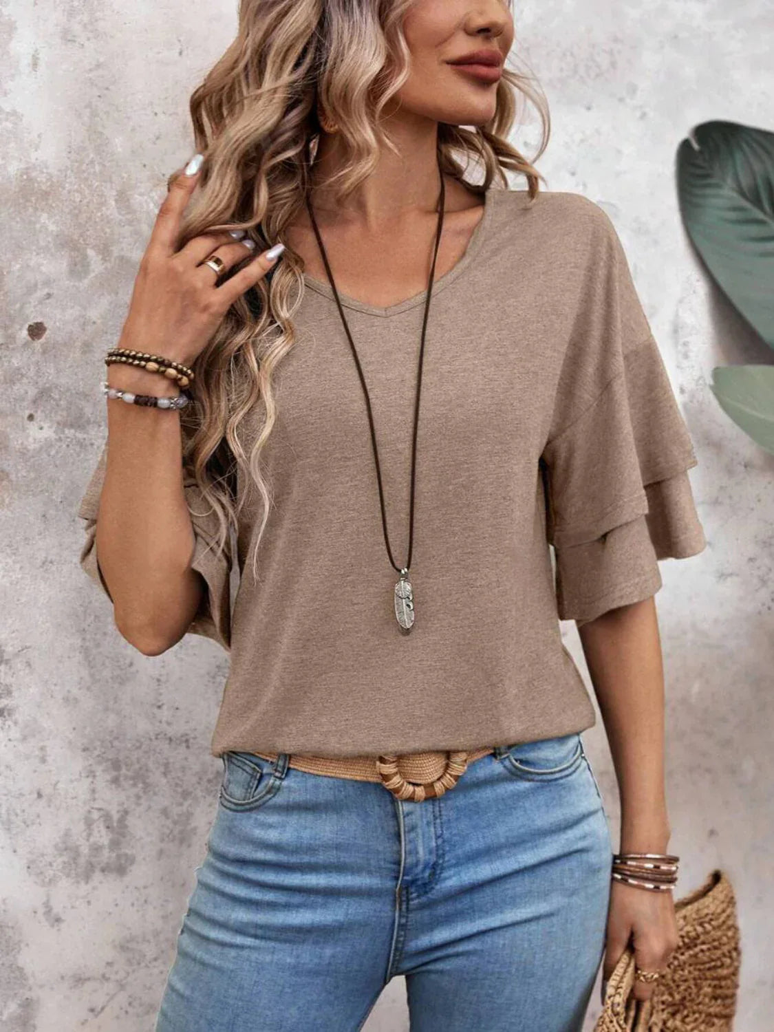 Woman in casual beige top with layered sleeves, wearing long necklace and blue jeans, standing against textured wall. Fashionable boho-chic style outfit.