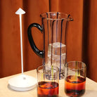 Elegant glass pitcher with ice cubes, two glasses of iced tea, and a modern white table lamp on a beige surface, set against a warm brown curtain backdrop.