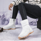 White winter snow boots with tan soles, worn by a person in black jeans and a striped sweater, set against a snowy mountain backdrop.