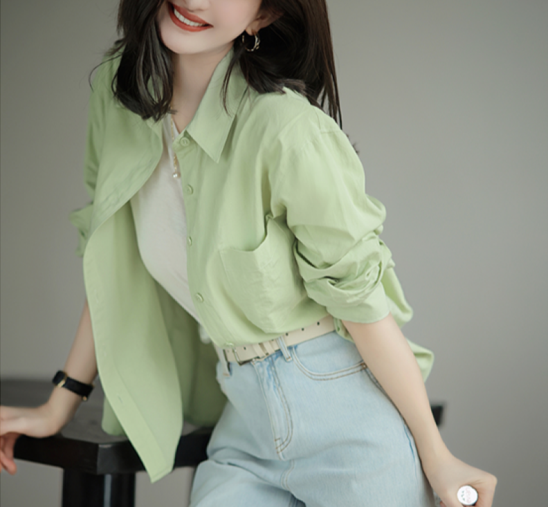 Woman in light green button-up shirt and light blue jeans, smiling, leaning on a table. Fashionable casual outfit, stylish women's clothing.