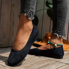 Woman wearing stylish black knit ballet flats with distressed jeans and turquoise anklet, standing on wooden deck. Fashionable casual footwear.