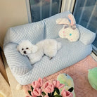 White fluffy dog resting on a light blue pet sofa with a plush bunny toy, near a window. Cozy pet furniture, cute dog, home decor.