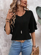 Woman in casual black top and blue jeans, accessorized with a long necklace and bracelets, standing against a textured wall. Fashionable casual outfit.