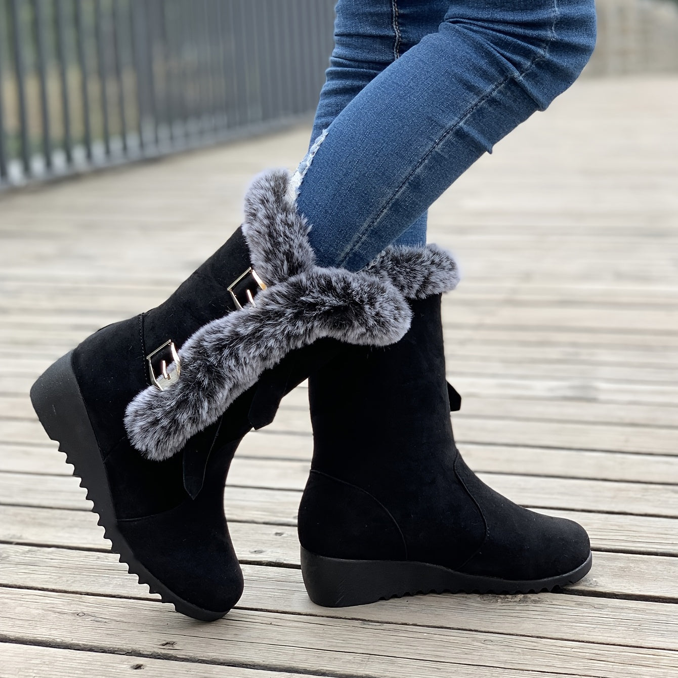 Black suede winter boots with faux fur trim and wedge heel, worn with blue jeans on a wooden deck. Stylish women's footwear for cold weather.