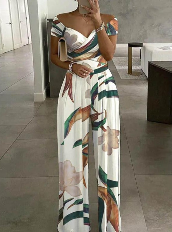 Woman wearing a stylish off-shoulder floral jumpsuit with wide-leg pants, holding a clutch in a modern, minimalist interior. Fashionable summer outfit.