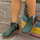 Green leather ankle boots with side zippers and brown soles, worn outdoors on colorful tire steps. Stylish women's footwear, casual fashion.