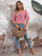 Woman in pink blouse and jeans holding a woven straw handbag, standing against a rustic wall with a tropical leaf. Fashionable casual outfit.