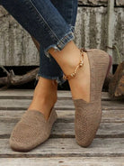 Beige knitted loafers on woman's feet, paired with distressed jeans and a gold anklet, on rustic wooden floor. Casual footwear fashion.