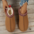 Brown fleece-lined winter slippers with red embroidered trim, worn by a person on wooden floor. Cozy, stylish footwear for cold weather comfort.