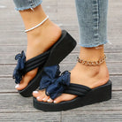 Women's black platform sandals with large bow detail, worn with anklets and jeans. Stylish summer footwear, trendy fashion accessory.