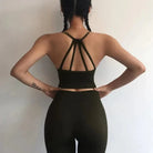 Woman wearing stylish black sports set with decorative back straps, braided hair, and tattoo on arm, standing against a plain background.