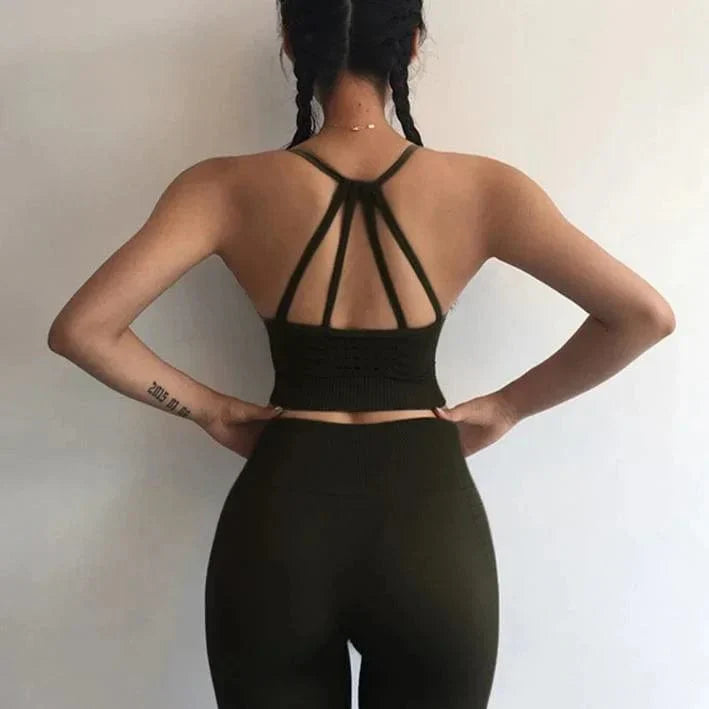 Woman wearing stylish black sports set with decorative back straps, braided hair, and tattoo on arm, standing against a plain background.