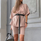 Woman wearing beige sports set with black belt, short-sleeve top, and biker shorts, standing indoors. Fashionable activewear outfit.