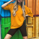 Woman in orange oversized t-shirt and black cycling shorts posing against colorful container backdrop. Fashionable casual outfit, street style.