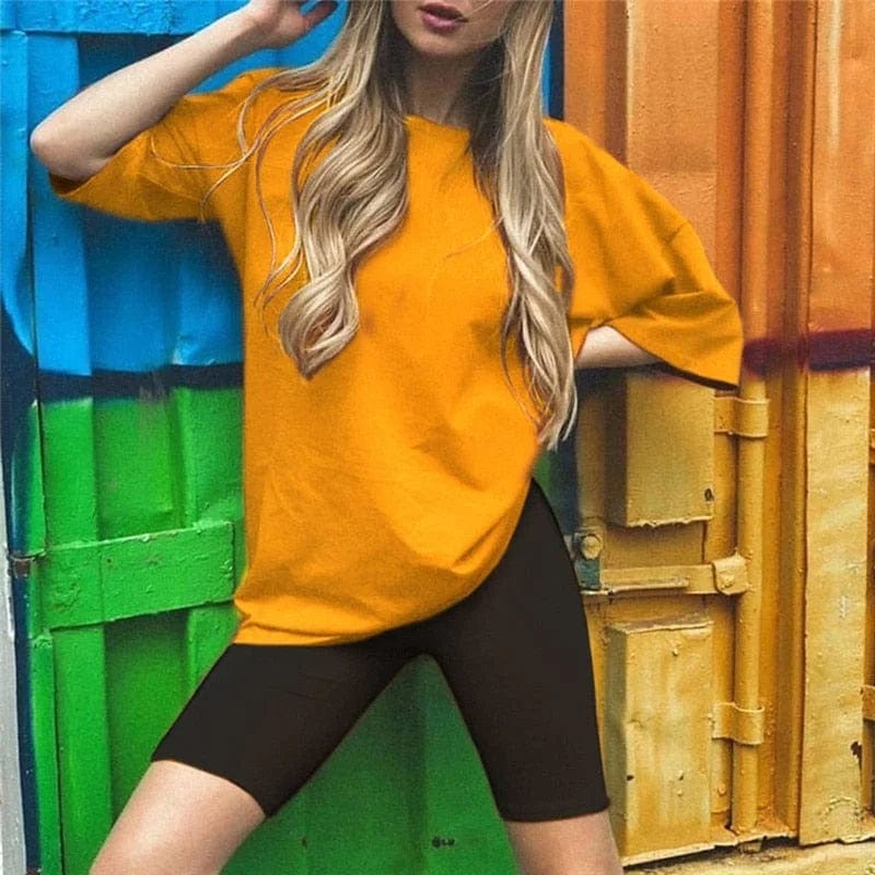 Woman in orange oversized t-shirt and black cycling shorts posing against colorful container backdrop. Fashionable casual outfit, street style.