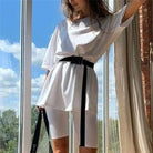 Woman in white oversized t-shirt and biker shorts with black belt, standing by a window. Casual summer outfit, trendy women's fashion.