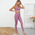 Woman wearing a pink sports bra and leggings set, standing in a modern room with wooden flooring and white panel backdrop.