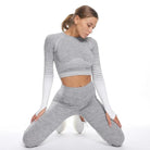 Woman wearing gray ombre-effect sports set, long-sleeve crop top, and high-waisted leggings, posing in a kneeling position on a white background.