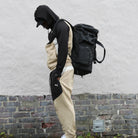 Man in beige and black tracksuit with hood, wearing a large black backpack, standing against a brick wall. Urban fashion, streetwear style.