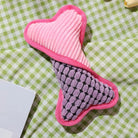 Pink and purple plush dog toy on green checkered fabric. Durable, textured, bone-shaped pet toy for chewing and play. Ideal for small to medium dogs.