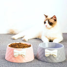 Pink and gray pet bowls with bow designs, filled with cat food, on a gray surface. Fluffy cat in the background. Ideal for pet feeding solutions.