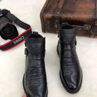 Black leather ankle boots with embossed design, placed on a white surface next to a vintage suitcase and Canon camera. Fashion footwear display.