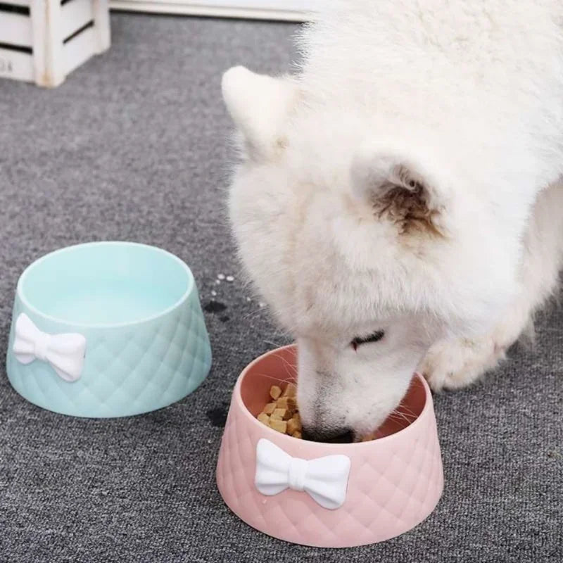 White dog eating from a pink, quilted dog bowl with a bow design, next to a blue bowl. Pet feeding accessories, stylish dog bowls, cute pet supplies.