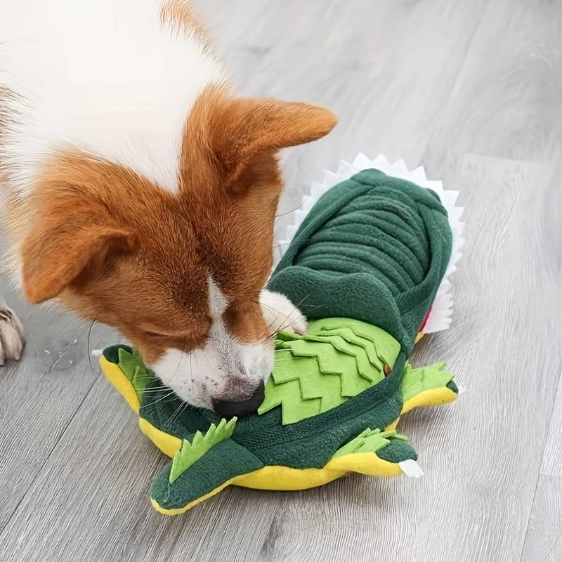 Dog playing with a green crocodile-shaped plush toy on a wooden floor. Durable pet chew toy, interactive dog toy, fun pet accessory.