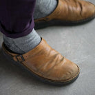 Brown leather clogs with adjustable strap, worn with gray socks and dark pants, on a concrete floor. Comfortable, casual footwear for everyday use.