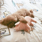 Fluffy brown dog cuddling a pink pig plush toy on a cartoon-patterned bedspread. Cozy pet scene, cute dog and toy, playful bedroom decor.