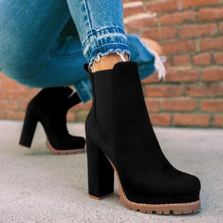 Black suede high-heeled ankle boots with chunky soles, worn with distressed blue jeans, against a brick wall background. Fashion footwear.