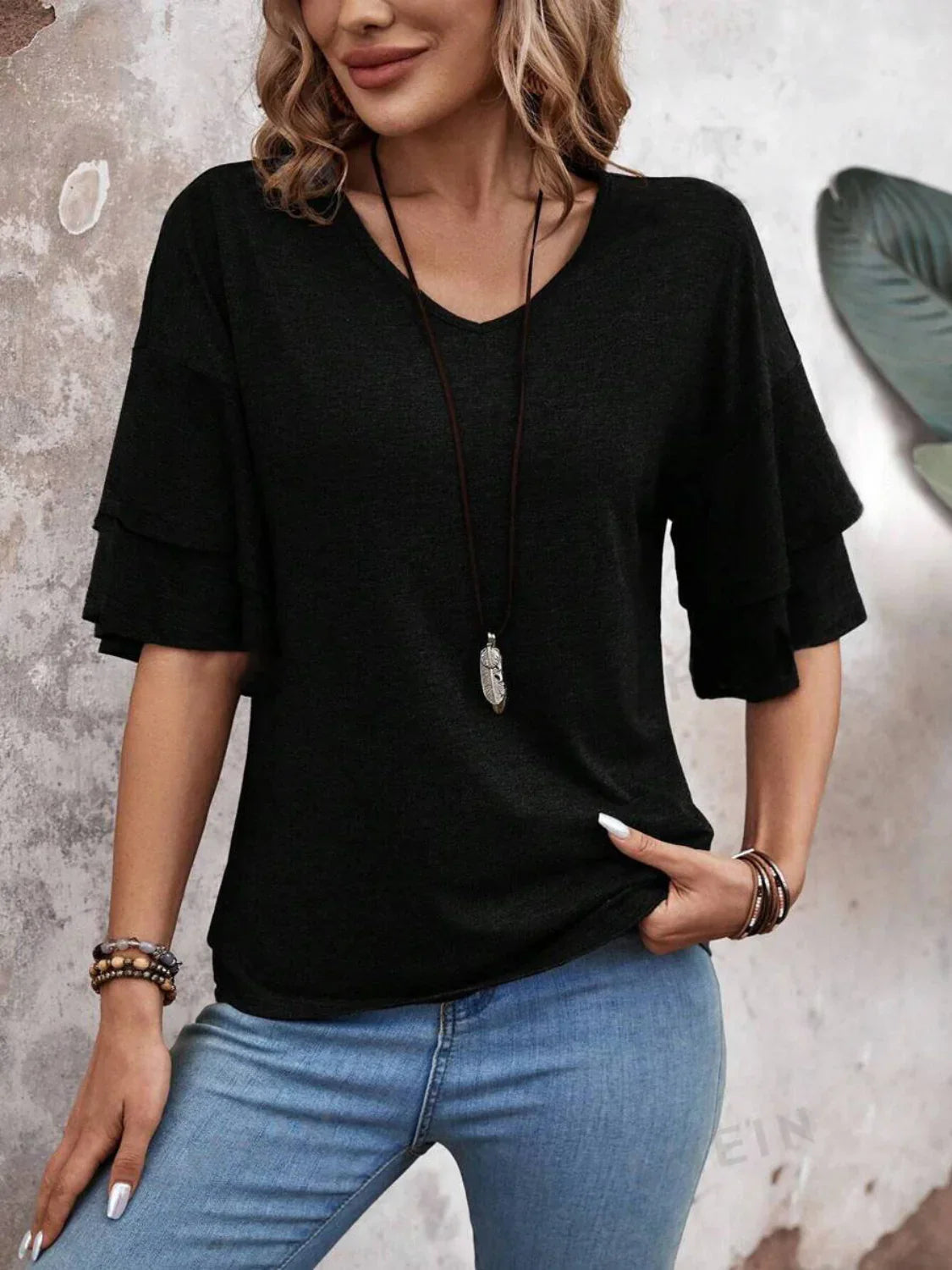 Woman wearing a black ruffle sleeve top and blue jeans, accessorized with a long necklace and bracelets, standing against a textured wall. Fashionable casual outfit.