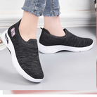 Black slip-on sneakers with white soles, worn with blue jeans. Comfortable, casual footwear ideal for walking and everyday use. Fashionable and lightweight.