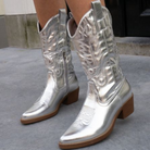 Silver cowboy boots with intricate stitching, mid-calf height, and wooden block heels on a concrete surface. Fashionable western footwear.